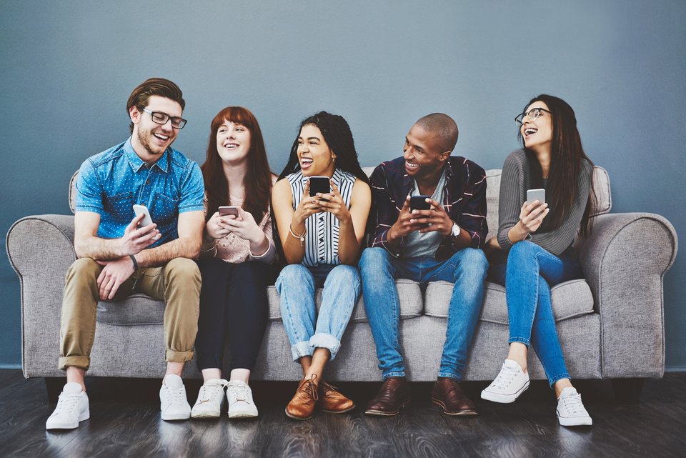 Eine Gruppe von jungen Menschen sitzen gut gelaunt und lachend, mit ihren Smartphones in den Händen, auf einem Sofa