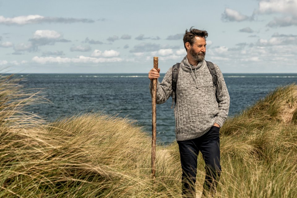 Ein Mann macht mit Pullover, Rucksack und Wanderstock eine Wanderung auf den Dünen, während hinter ihm das Meer zusehen ist, und versucht sich zu erholen und neue Kräfte zu sammeln