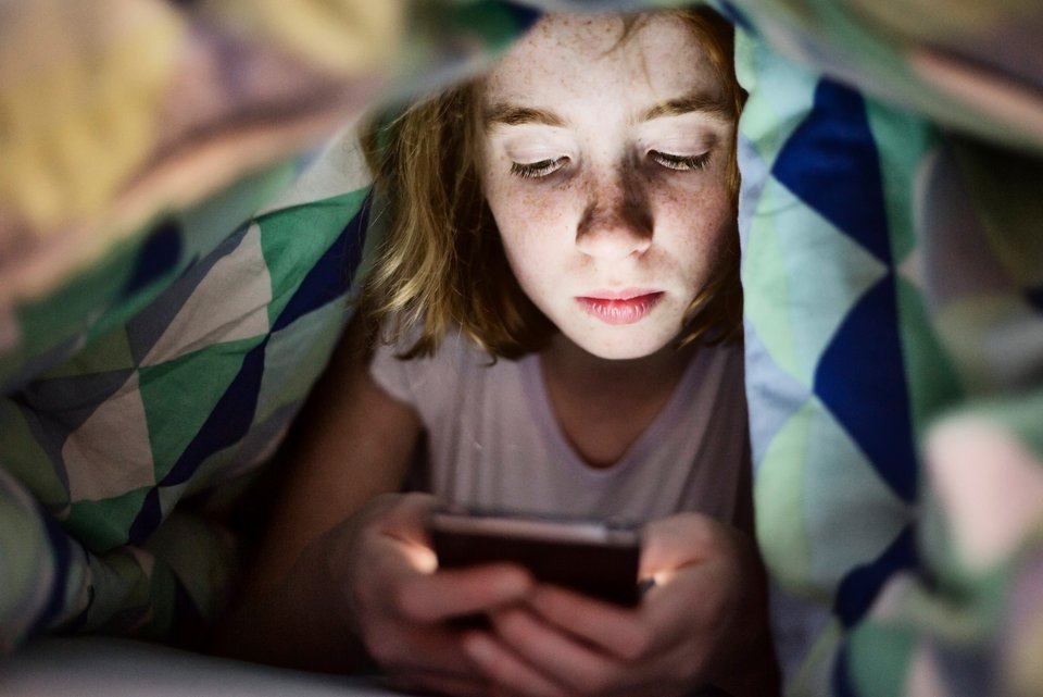 Ein Mädchen schaut konzentriert auf ihr hell erleuchtetes Smartphone-Display während es um sie herum dunkel ist. Sie versteckt sich dabei unter der Bettdecke.