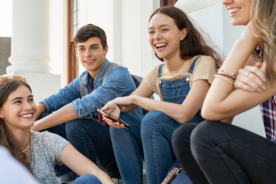 Eine Gruppe von pubertierenden Teenagern sitzen lachend auf einer Treppenstufe vor einem Gebäude