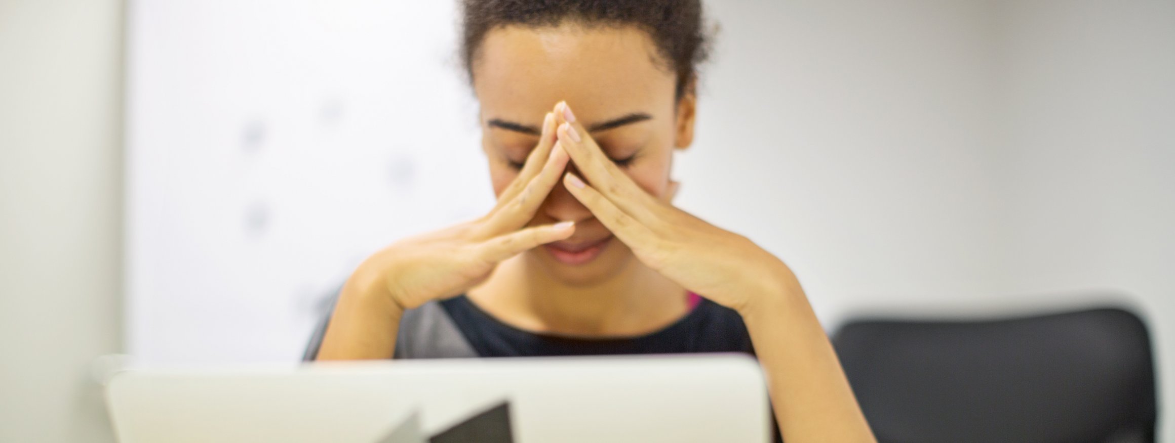 Eine junge Frau sitzt vor ihrem Laptop, die Hände vor das Gesicht gefaltet und wirkt müde