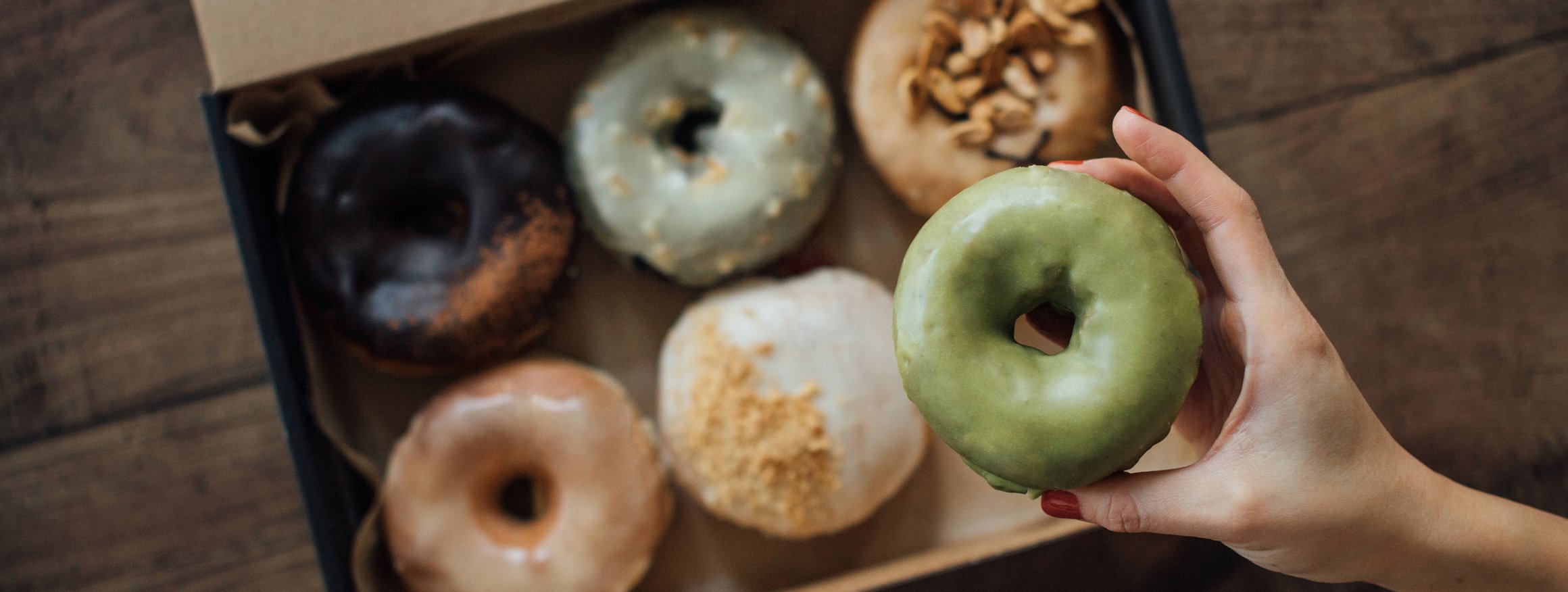 Eine Frauenhand greift in eine Donutschachtel und holt sich einem Donut mit grüner Glasur heraus