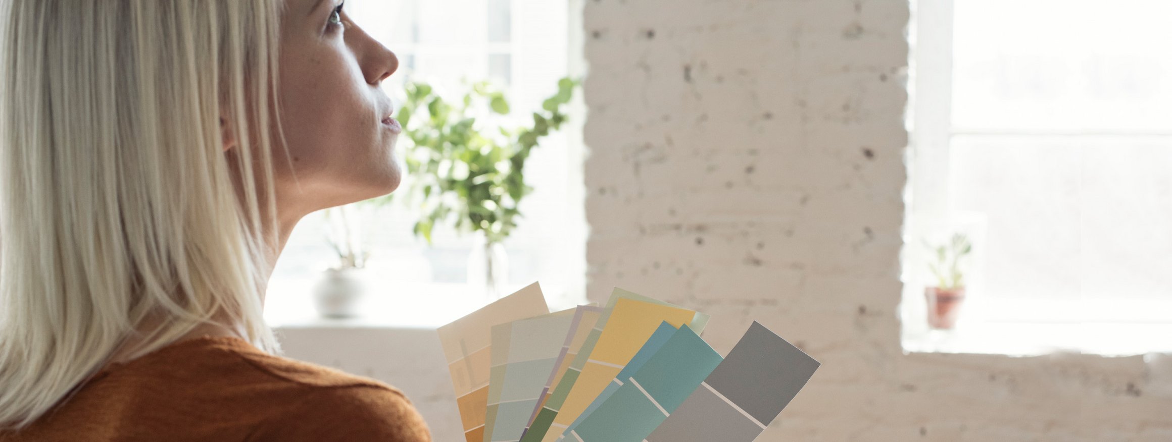 Eine junge blonde Frau steht in einem Raum mit einem Farbmustern in der Hand und überlegt, welche Wandfarbe sie nehmen soll