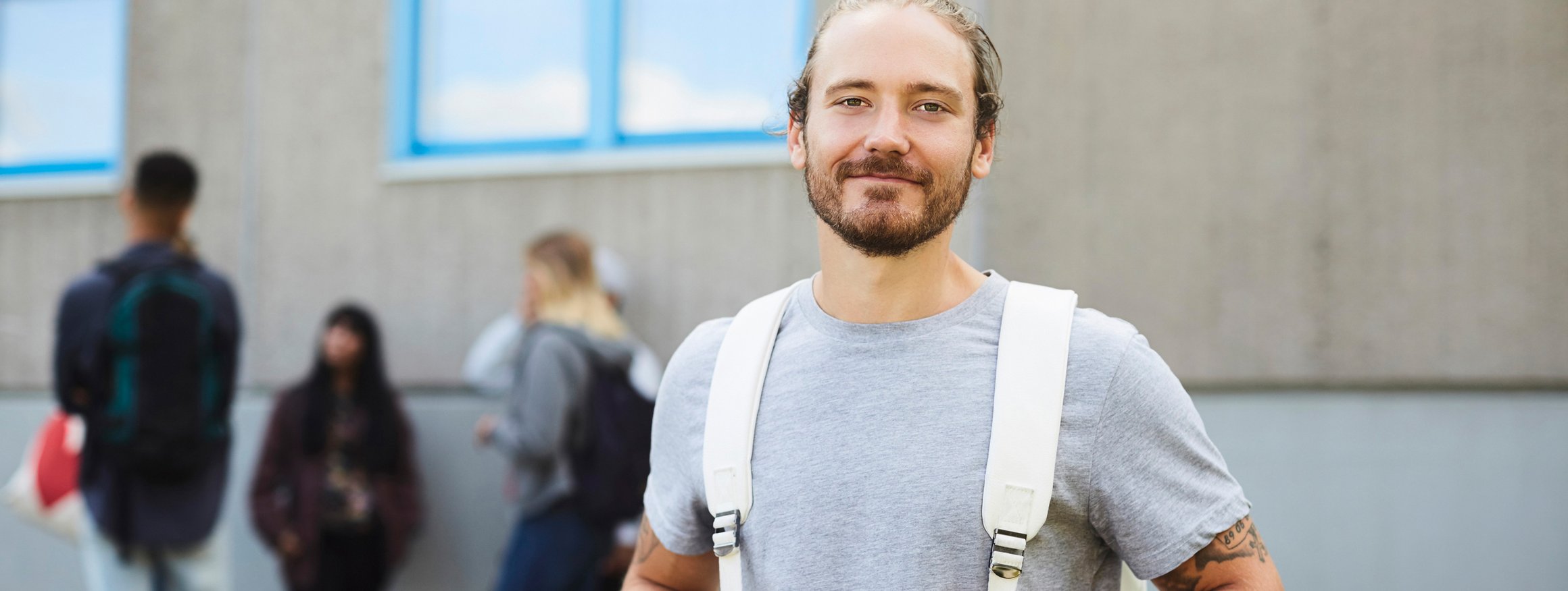 Ein blonder Student mit blondem Bart und Tattoo steht mit weißen Rucksack da, hinter ihm stehen seine Kommilitoninnen und Kommilitonen.