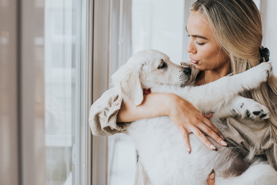 Eine junge Frau mit blonden langen Haaren hält fürsorglich einen jungen Hund im Arm und redet einfühlsam mit ihm