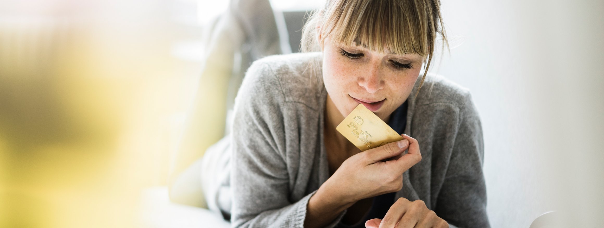 Eine junge Frau liegt auf dem Bauch auf einem Sofa, das Smartphone vor ihr und hält dabei ihre Kreditkarte in der Hand, da sie dem Kaufangebot nicht widerstehen kann