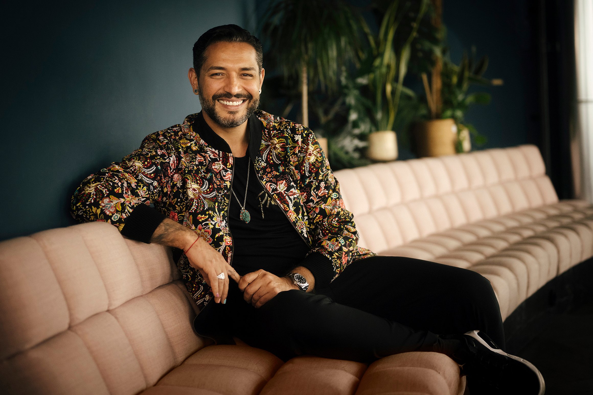 Das Foto zeigt Gianni Jovanovic, wie er auf einem hellen Sofa sitzt. Der Mann lächelt freundlich.