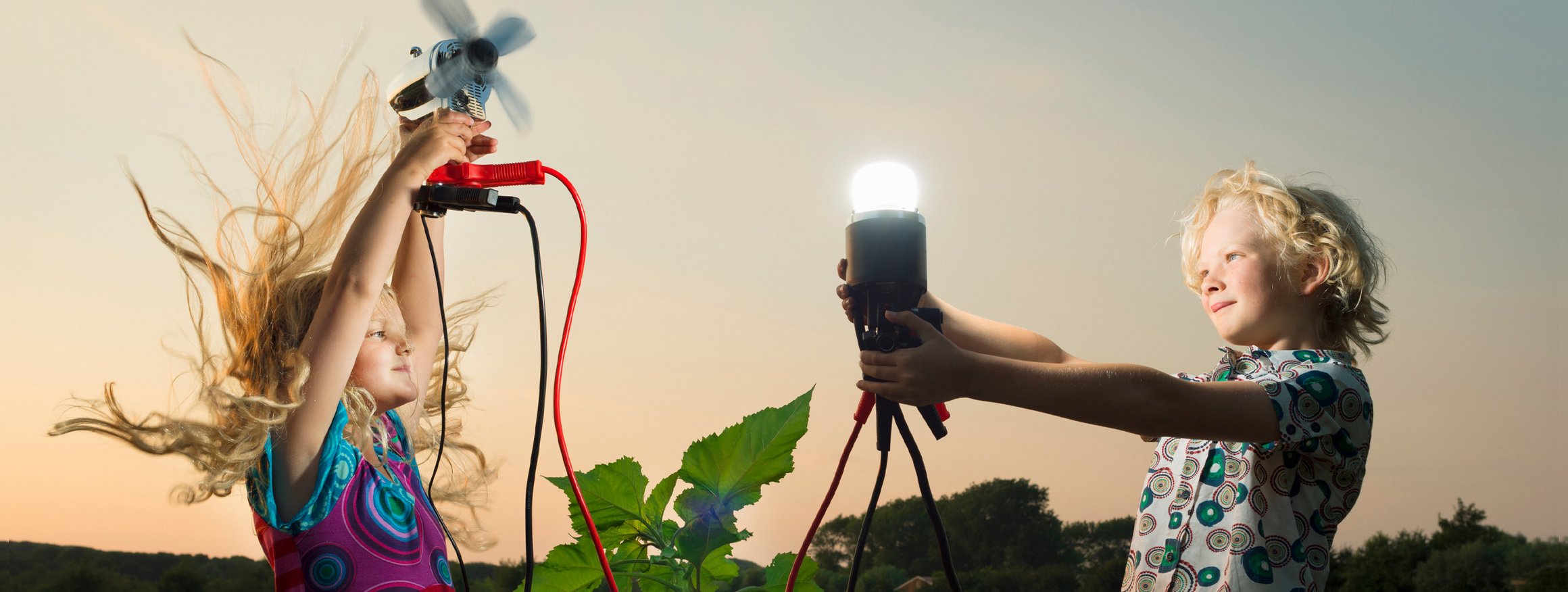 Zwei Kinder stehen an einem Feld mit Sonnenblumen und halten jeweils eine Lampe und einen Ventilator in den Händen, während sich bei dem Mädchen die Haare durch den Luftzug des Ventilators hochstellen