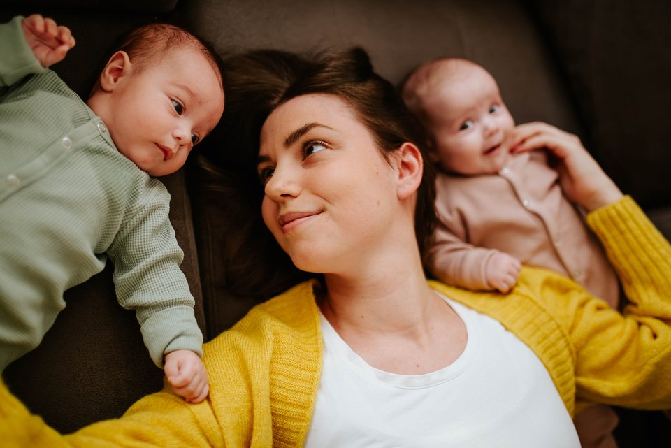 Eíne junge Mutter liegt zusammen mit ihren beiden Babies da und berührt sie sanft und schaut dabei liebevoll