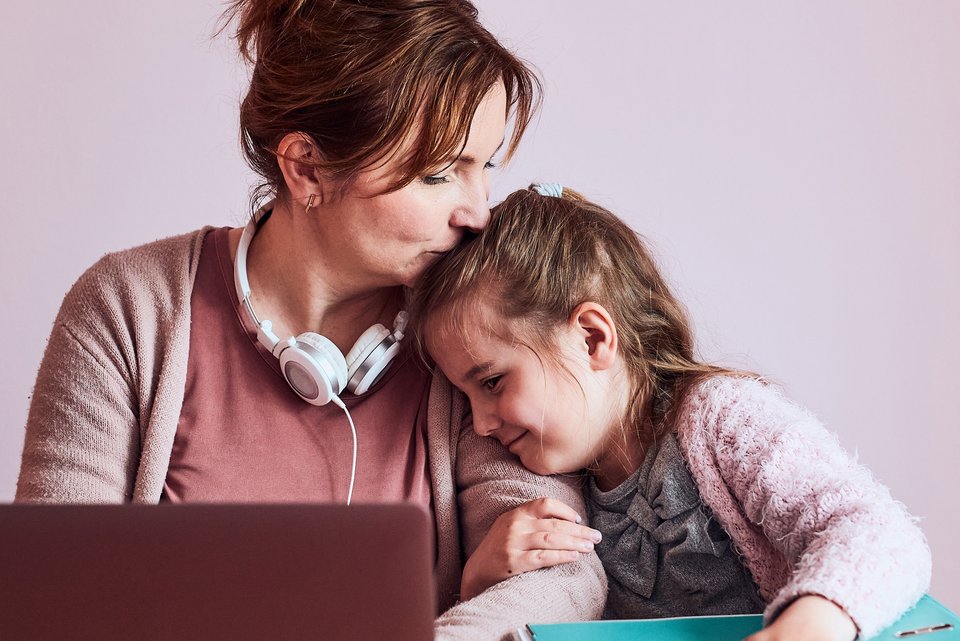 Eine alleinerziehende Mutter sitzt vor ihrem Computer im Homeoffice, daneben ihre kleine Tochter, die sich an die Mutter lehnt, die ihr fürsorglich den Kopf küsst