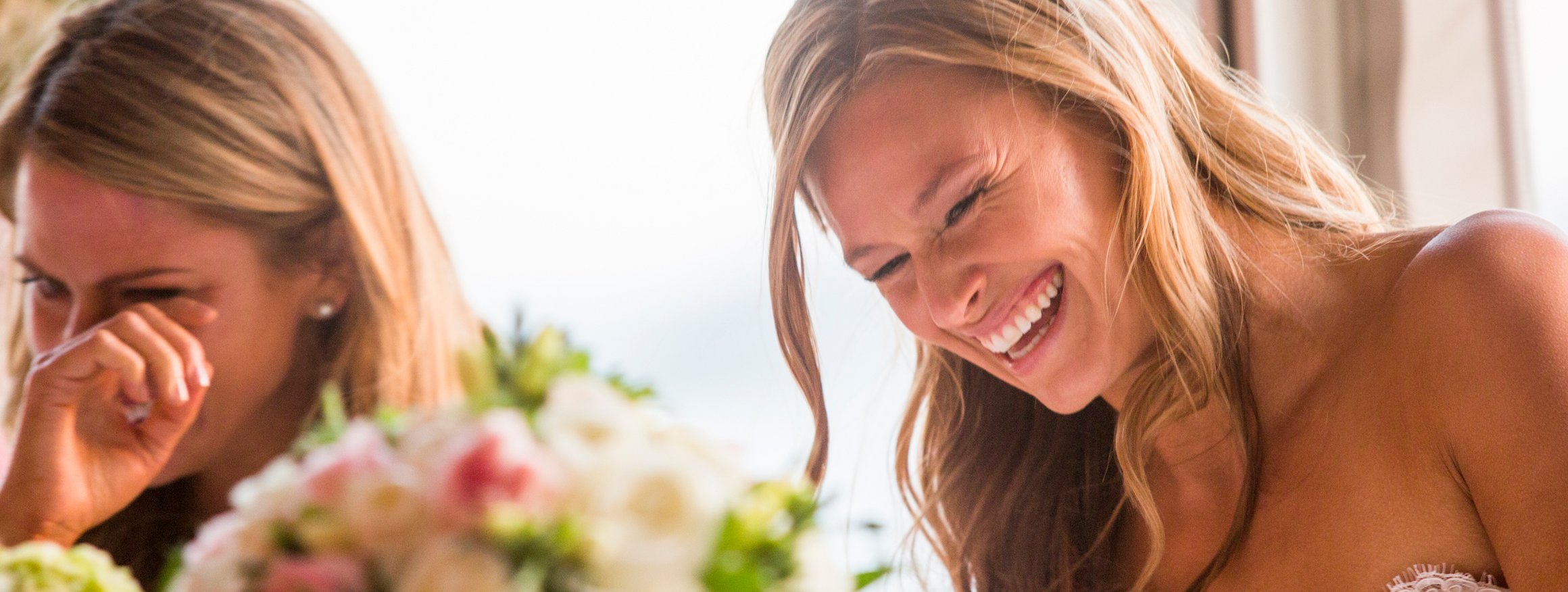 Braut im Brautkleid an einem Tisch mit Blumengestecken, lacht glücklich und ein weiblicher Hochzeitsgast wischt sich vor Rührung eine Träne aus dem Auge