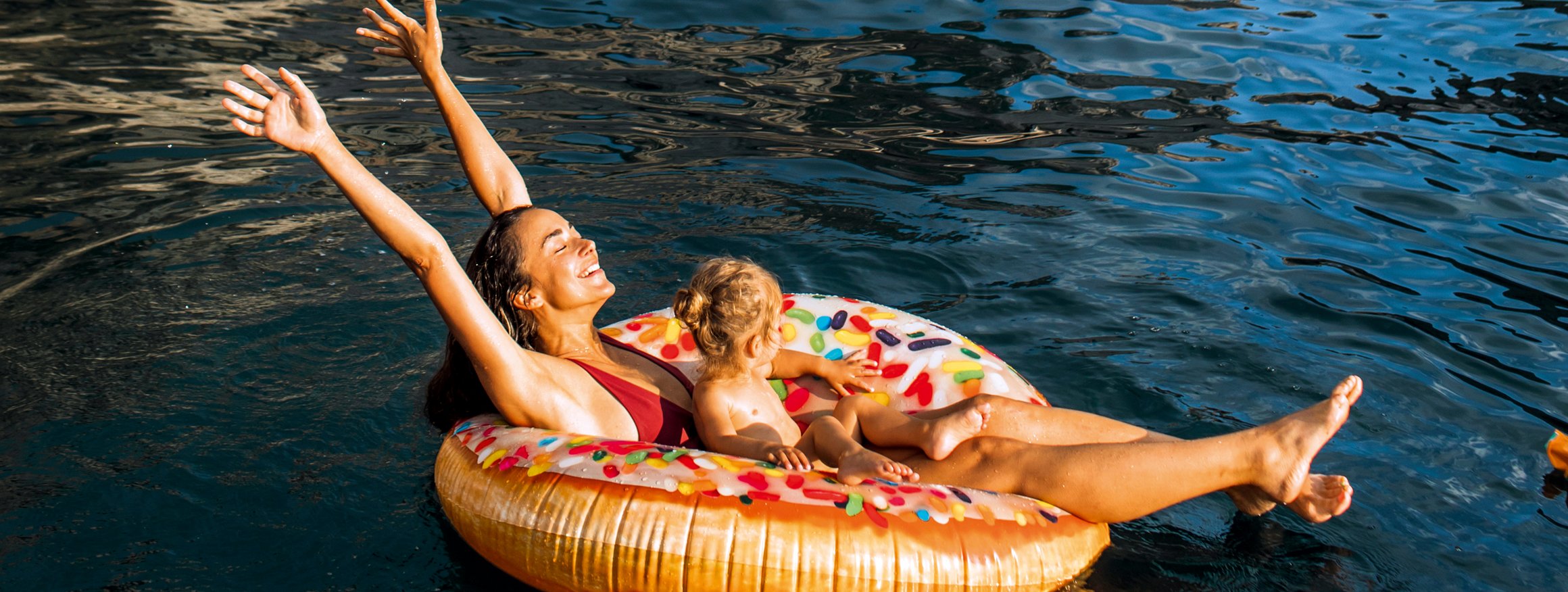 Eine Frau sitzt mit Badekleidung und ihrem Kind auf einer Schwimminsel im Wasser und reckt lachend die Arme nach oben