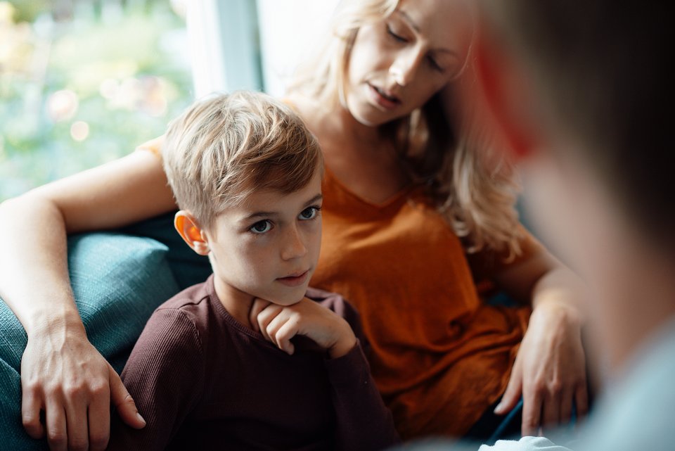 Ein Junge sitzt bei seiner Mutter, die ihren Arm um das Kind gelegt hat, und der seinem Vater anschaut, der vor ihm sitzt 