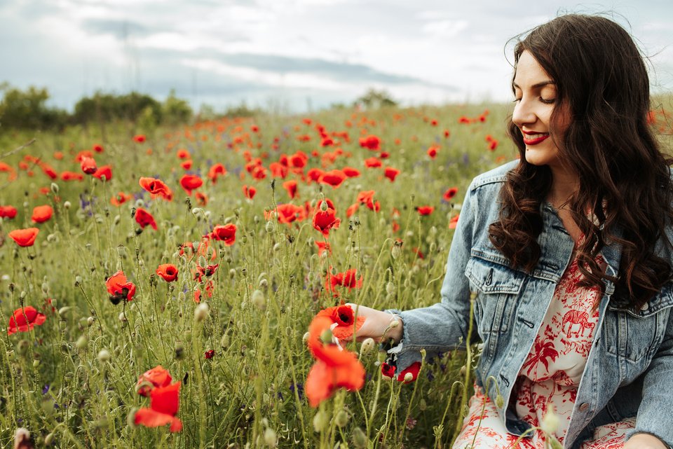 Eine junge Frau in Jeansjacke mit rotem Lippenstift und langen dunklen Haaren sitzt lächelnd in einem roten Mohnblumenfeld