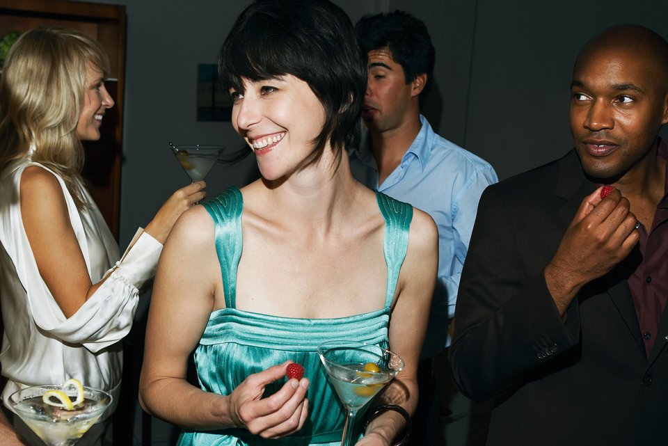 Eine junge Frau steht mit einem Cocktail lachend in einer Gruppe von Menschen auf einer Party