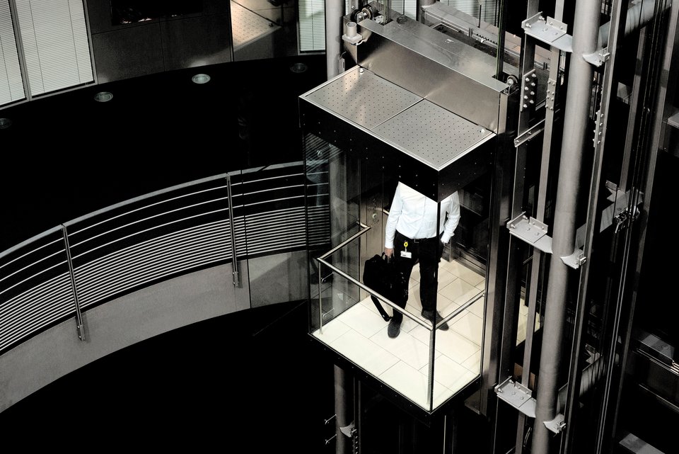 Ein Mann im Business-Look steht in einem gläsernen Aufzug in einem sehr modernen, aber kalten Bürogebäude
