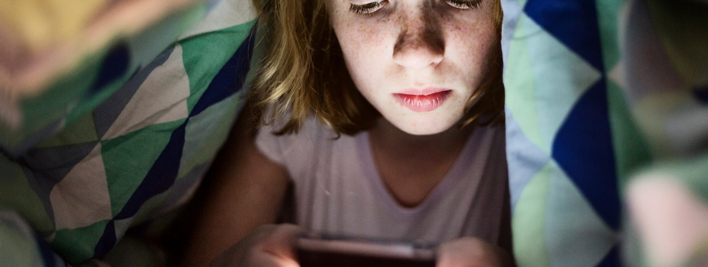 Ein Mädchen schaut konzentriert auf ihr hell erleuchtetes Smartphone-Display während es um sie herum dunkel ist. Sie versteckt sich dabei unter der Bettdecke.