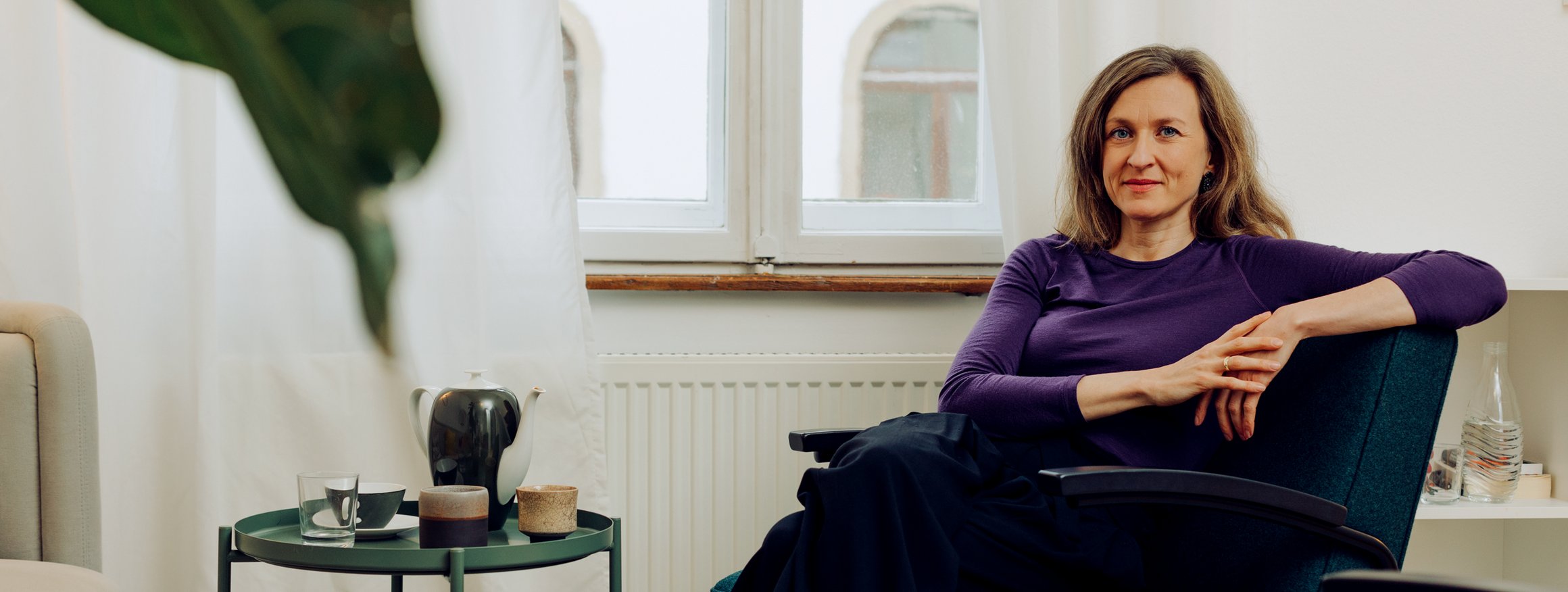 Die Therapeutin Angelika Eck sitzt ruhig lächelnd auf einem Sessel in ihrer Karlsruher Praxis, neben sich ein Tisch mit einer Teekanne, Tassen und einem Glas