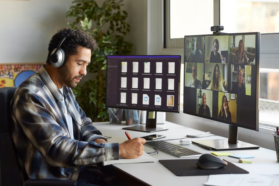 Ein Schwarzer Mann sitzt am Schreibtisch im Home-Office. Er trägt Kopfhörer und notiert etwas während eines digitalen Meetings.