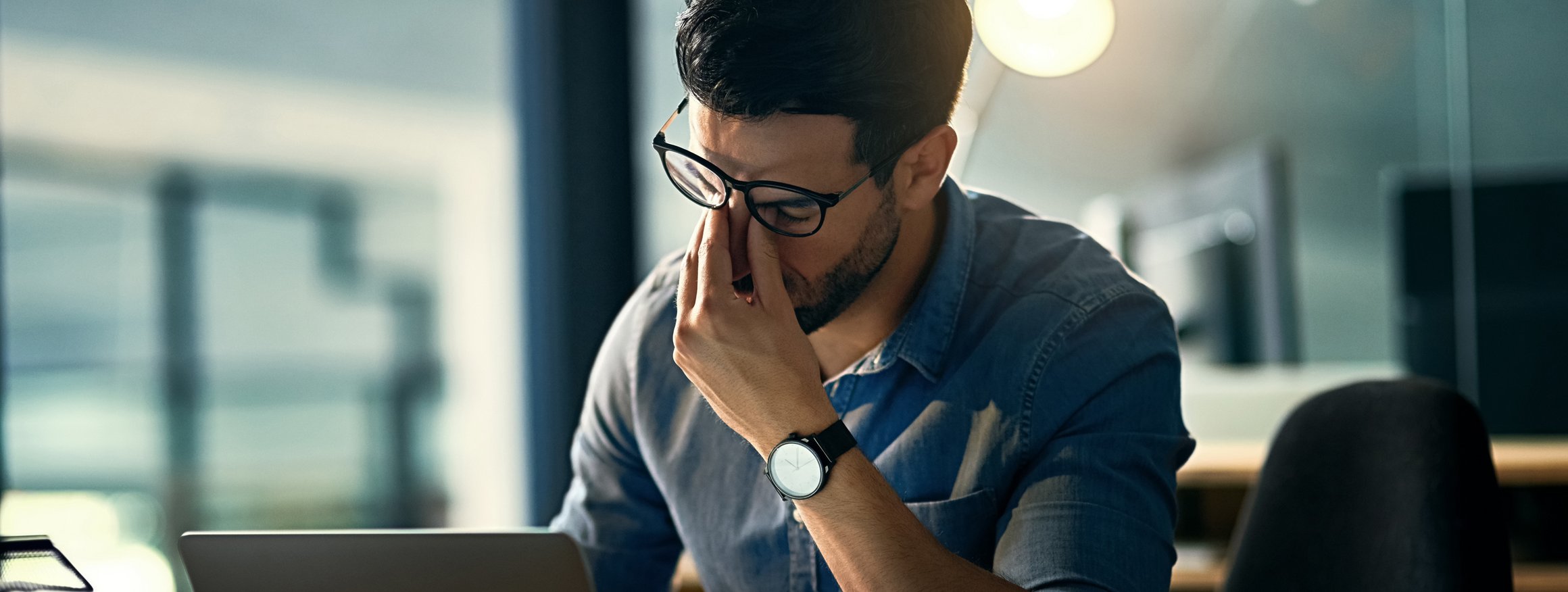 Ein Mann sitzt gestresst an seinem Laptop und schiebt sich die Brille hoch, um sich die müden Augen zu reiben