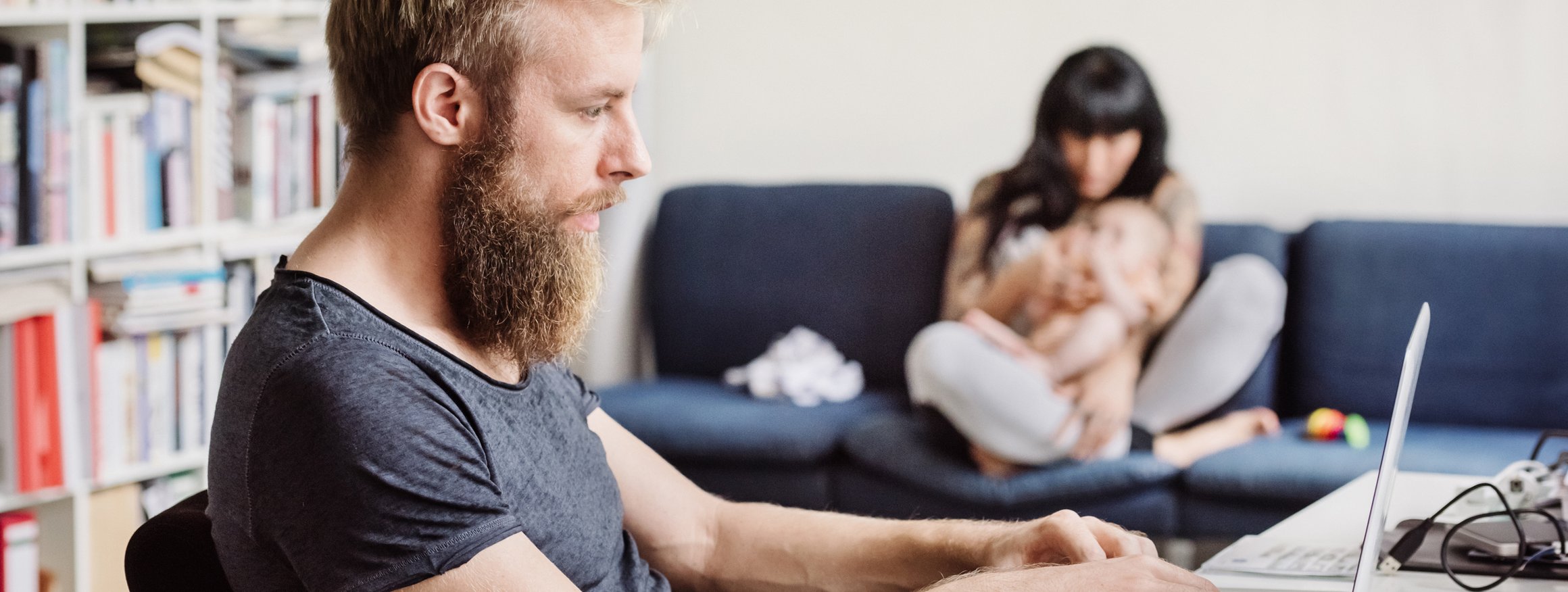 Ein jüngerer Mann sitzt in seinem Wohnzimmer im Homeoffice, dahinter sitzt seine Partnerin mit einen Baby auf dem Arm