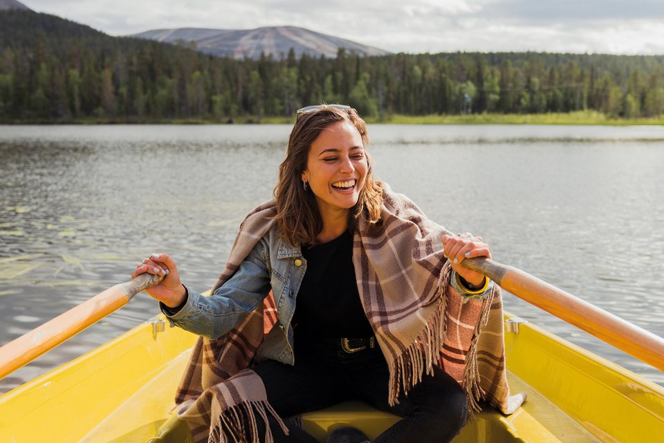Eine junge Finnin sitzt zufrieden lachend in einem gelben Ruderboot und rudert auf einem See