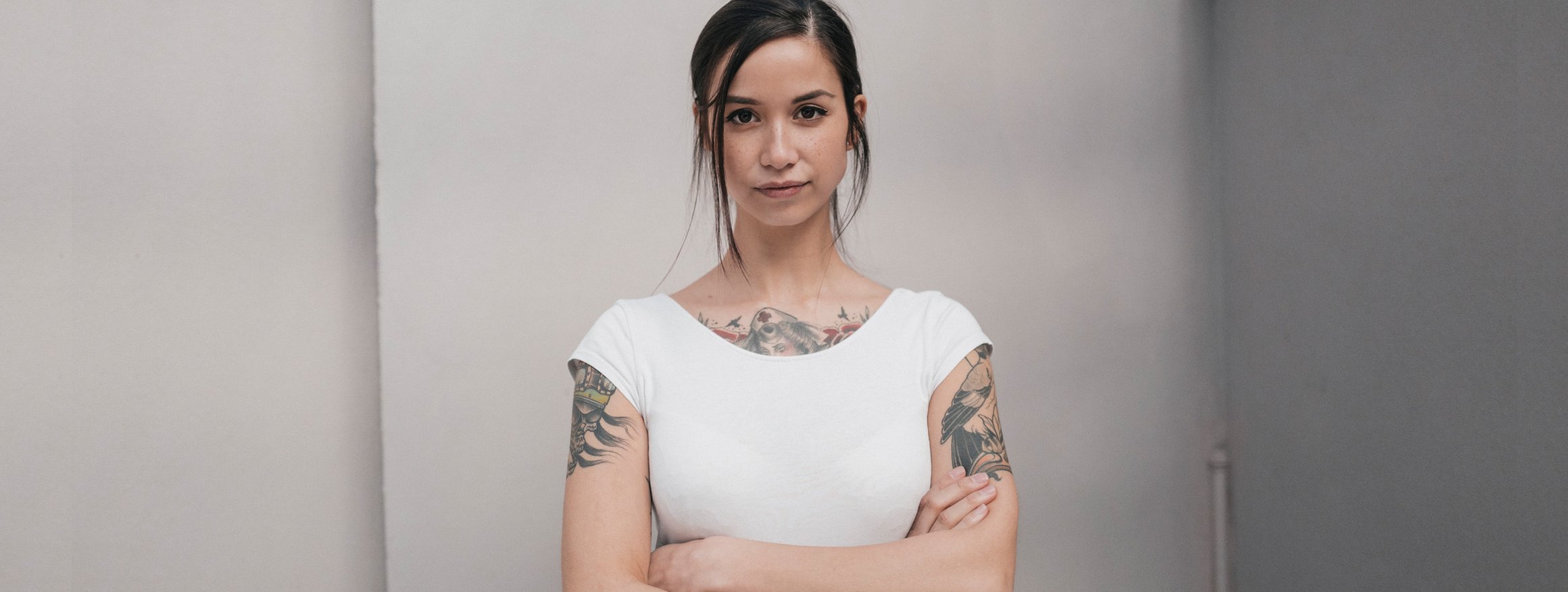 Eine junge Frau mit weißem Shirt und Tattoos schaut ernst und hat die Arme vor ihren Körper verschränkt