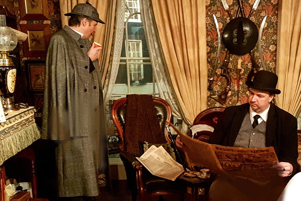 Sherlock Holmes steht in einem Salon, daneben sitzt Dr. Watson und liest in der Zeitung