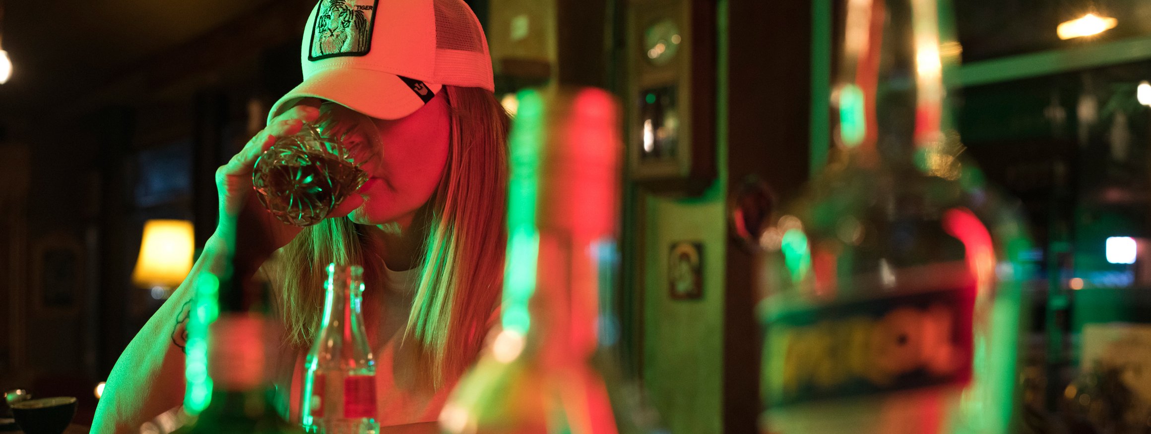 Eine Frau mit Basecap sitzt an einer Theke mit gedimmten Licht und trinkt, während hinter ihr viele Flaschen im Regal stehen mit alkoholischen Getränken