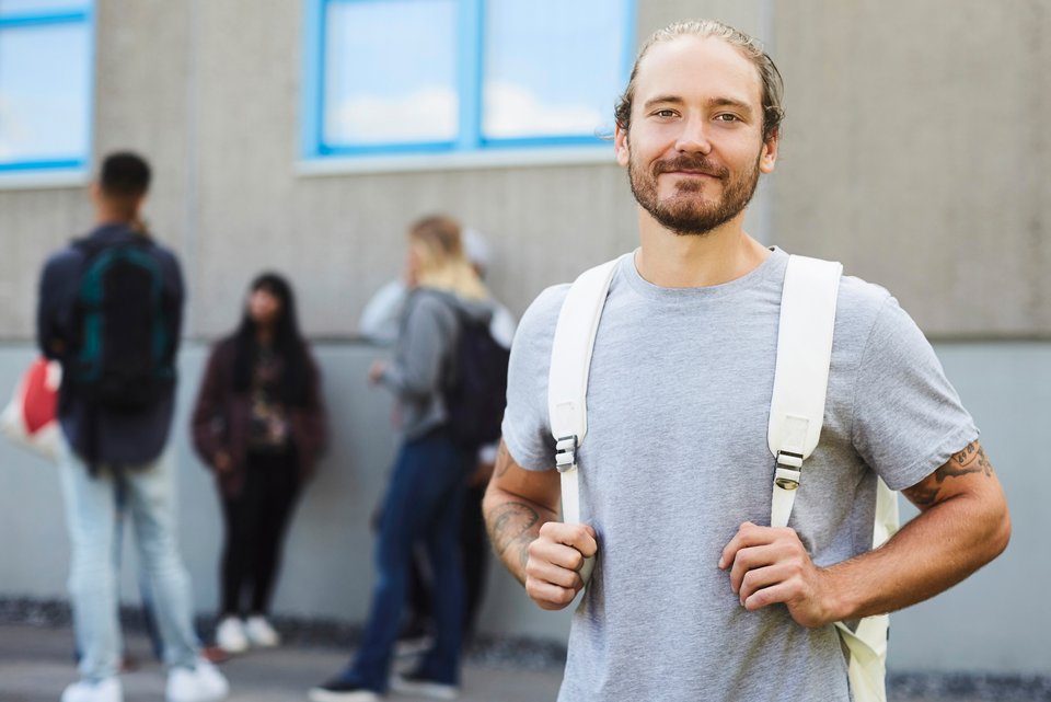 Ein blonder Student mit blondem Bart und Tattoo steht mit weißem Rucksack da, hinter ihm stehen seine Kommilitoninnen und Kommilitonen.