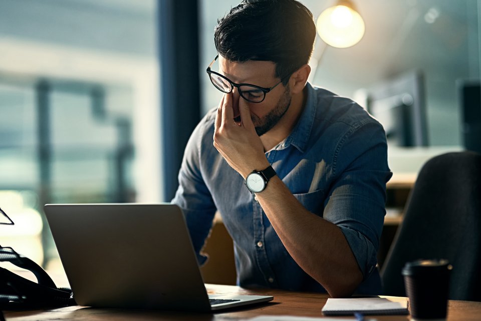 Ein Mann sitzt gestresst an seinem Laptop und schiebt sich die Brille hoch, um sich die müden Augen zu reiben