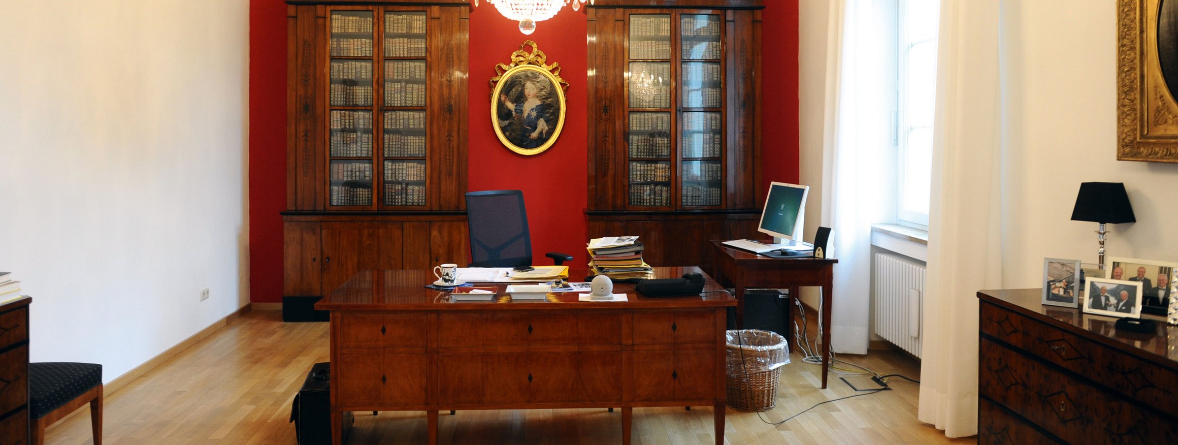 Das Bürozimmer einer Führungskraft mit antiken Schreibtisch, Aktenstapel, Bildschirm und einer Bibliothek im Hintergrund mit einem altertümlichen Gemälde an der rot gestrichenen Wand, an der Decke der Kronleuchter