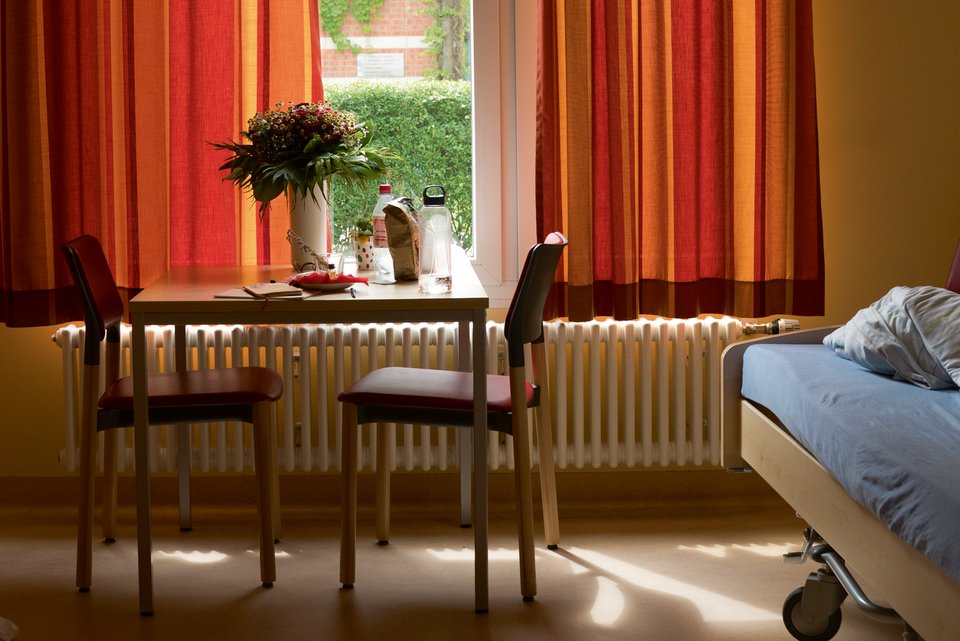Ein Zimmer in der Psychiatrie mit Bett, Tisch, zwei Stühlen und ein Blumenstrauß steht auf dem Tisch, während die Vorhänge am Fenster halb zugezogen sind