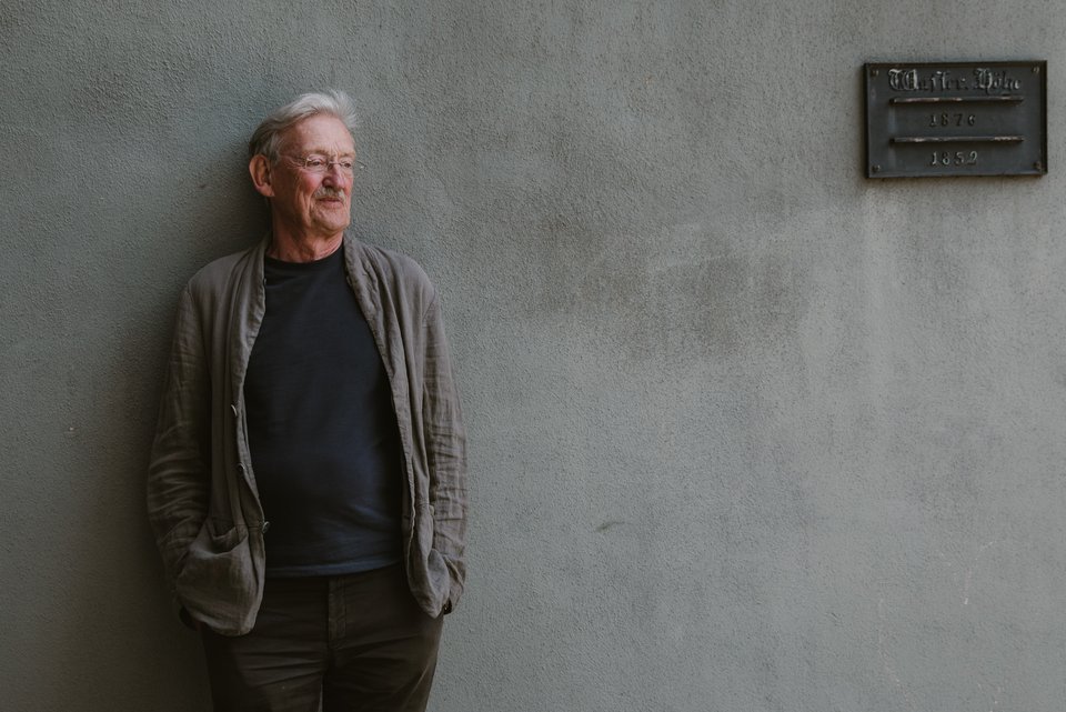 Der Schriftsteller, Christian Haller, lehnt an eine graue Wand mit einer Gedenktafel, die Hände in den Hosentaschen
