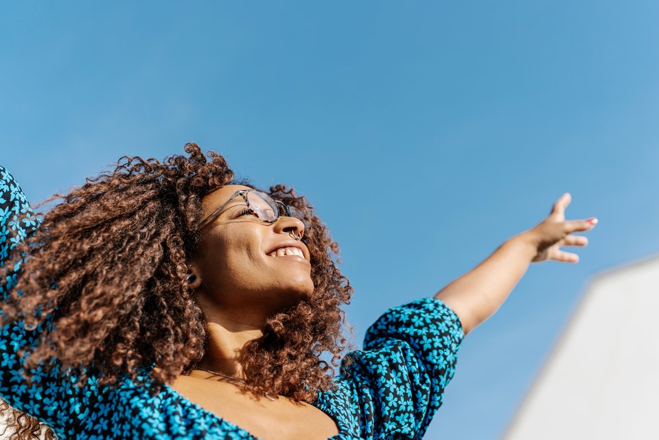 Eine junge Frau mit Locken reckt glücklich die Arme in den Himmel, weil sie glaubt, immer positiv zu denken fördert die Gesundheit