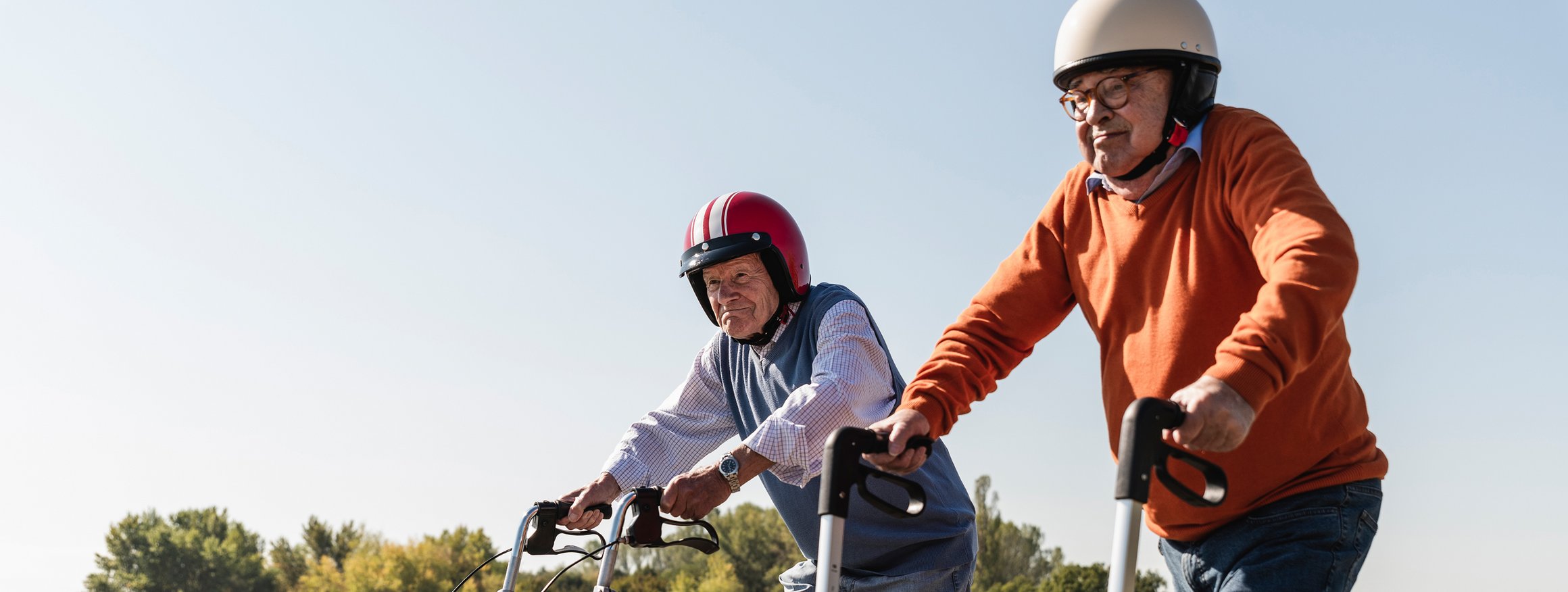 Zwei alte Männer tragen Helme und laufen vergnügt mit ihrem Rollatoren die Straße entlang