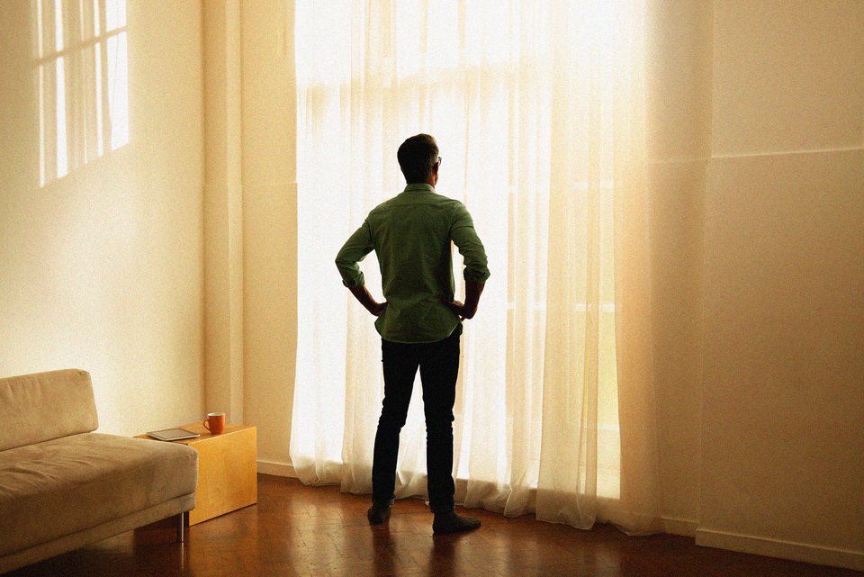 Ein älterer Mann steht traurig in einem Raum und schaut aus dem Fenster, die Hände an den Hüften