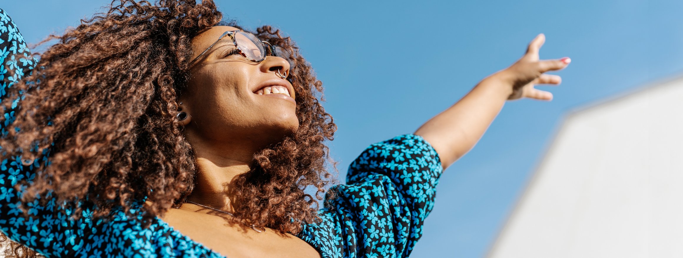 Eine junge Frau mit Locken reckt glücklich die Arme in den Himmel, weil sie glaubt, immer positiv zu denken fördert die Gesundheit