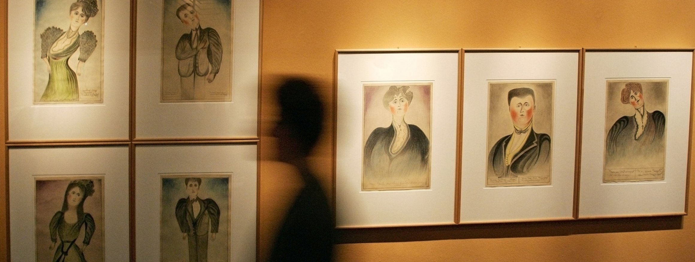 Das Foto zeigt eine menschliche Silhouette, die an Gemälden vorbeiläuft. Die Kunstwerke zeigen Selbstporträts von Psychiatrie-Erfahrenen.