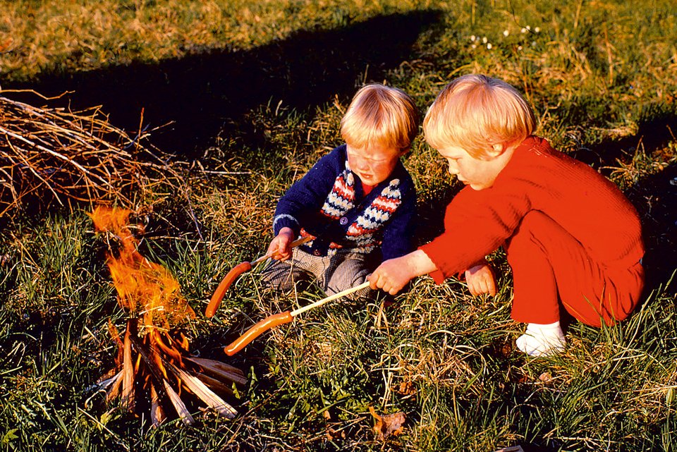 Ein älteres Foto zeigt zwei blonde Kinder auf einer Wiese mit Würstchen auf Stöcken gespießt vor einem Lagerfeuer