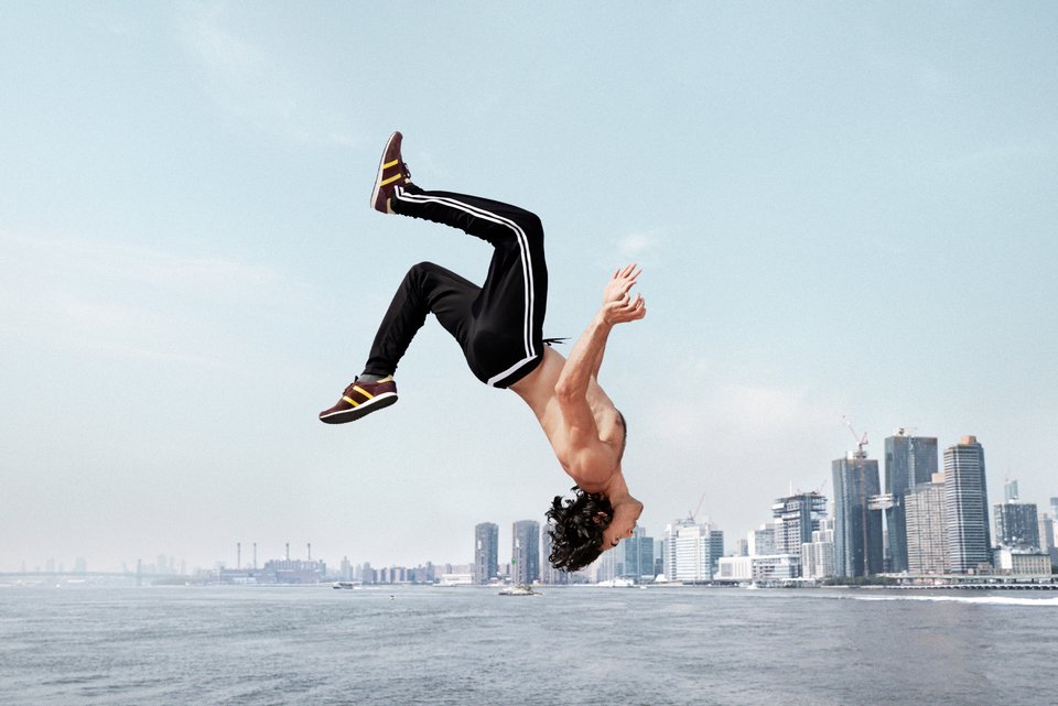 Ein Mann mit Sporthosen, Sportschuhen und nacktem Oberkörper macht einen riskanten Salto an einem Fluss, dahinter die Skyline einer großen Stadt