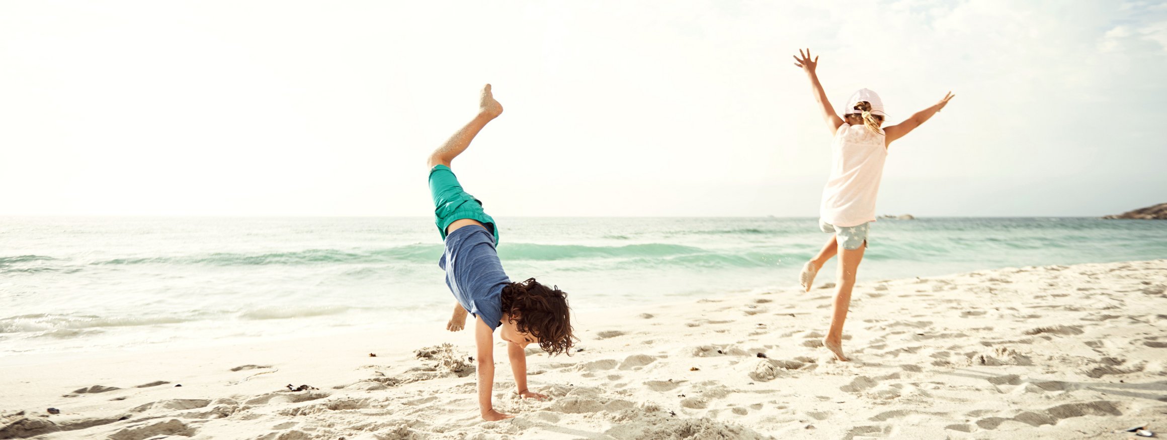 Zwei Kinder springen frei und ausgelassen an einem weißen Strand und der Junge schlägt dabei ein Rad