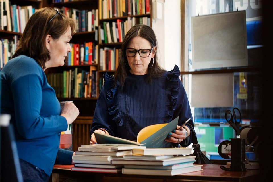 Zwei weibliche Bekannte stehen vor einer Bücherwand an einem Tisch mit vielen Bücher und unterhalten sich, während sie in einem Buch blättern