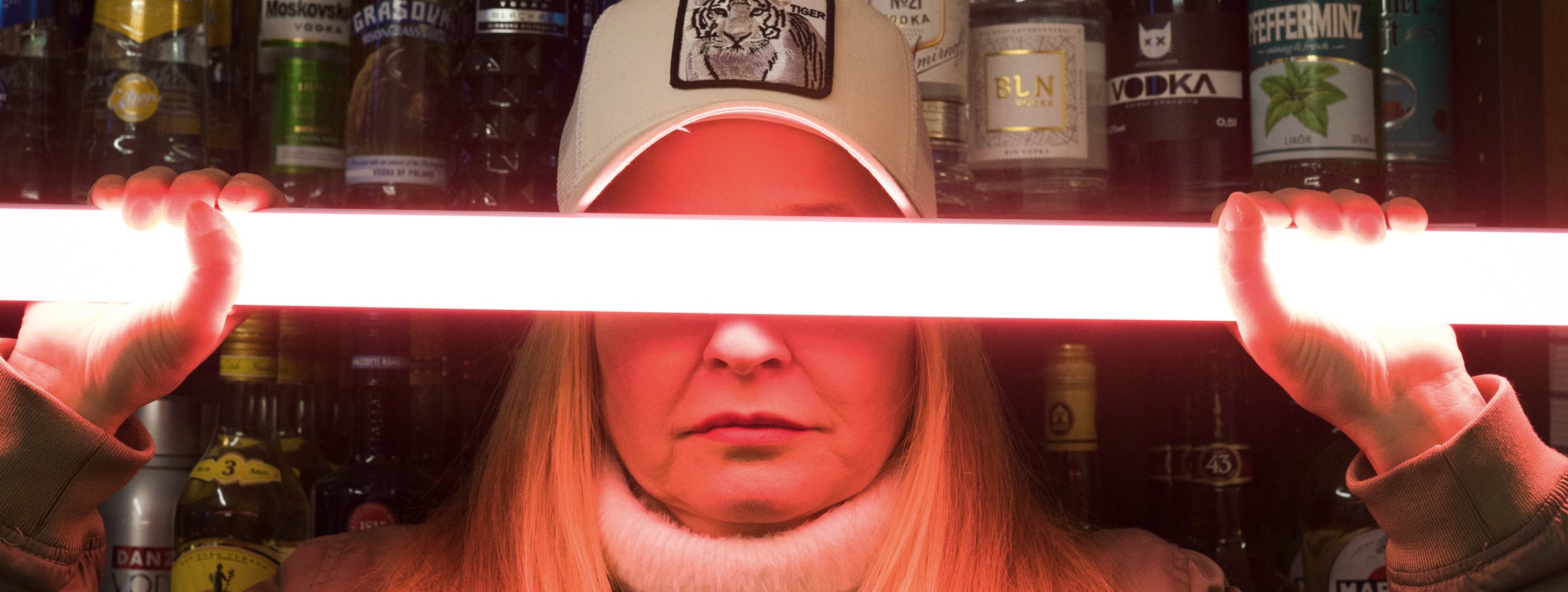 Eine Frau hält sich einen Leuchtstab vor die Augen, hinter ihr ist eine Thekenbar mit vielen alkoholischen Getränken in Flaschen