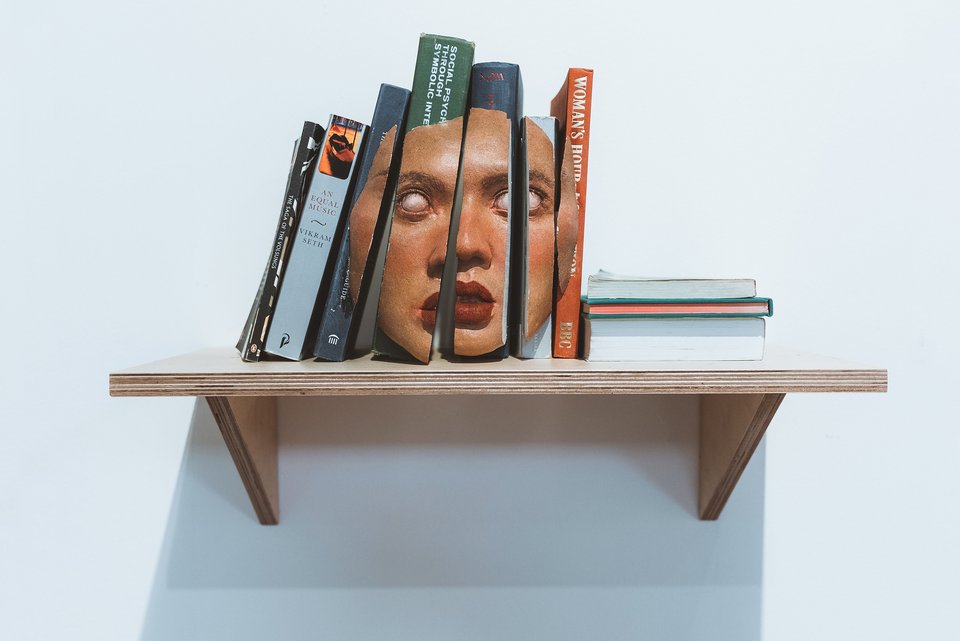 Das Foto zeigt mehrere Bücher, die auf einem Regalbrett stehen. Die Buchrücken ergeben zusammen eine Maske mit ausdruckslosem Blick.