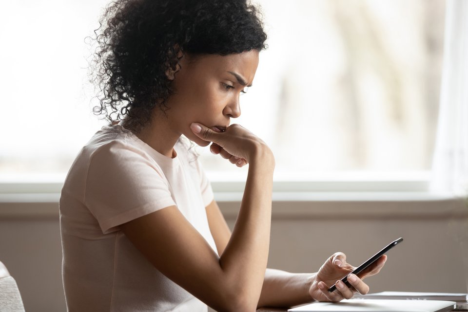 Eine junge Frau sitzt skeptisch und gestresst am Tisch und schaut dabei auf ein Smartphone