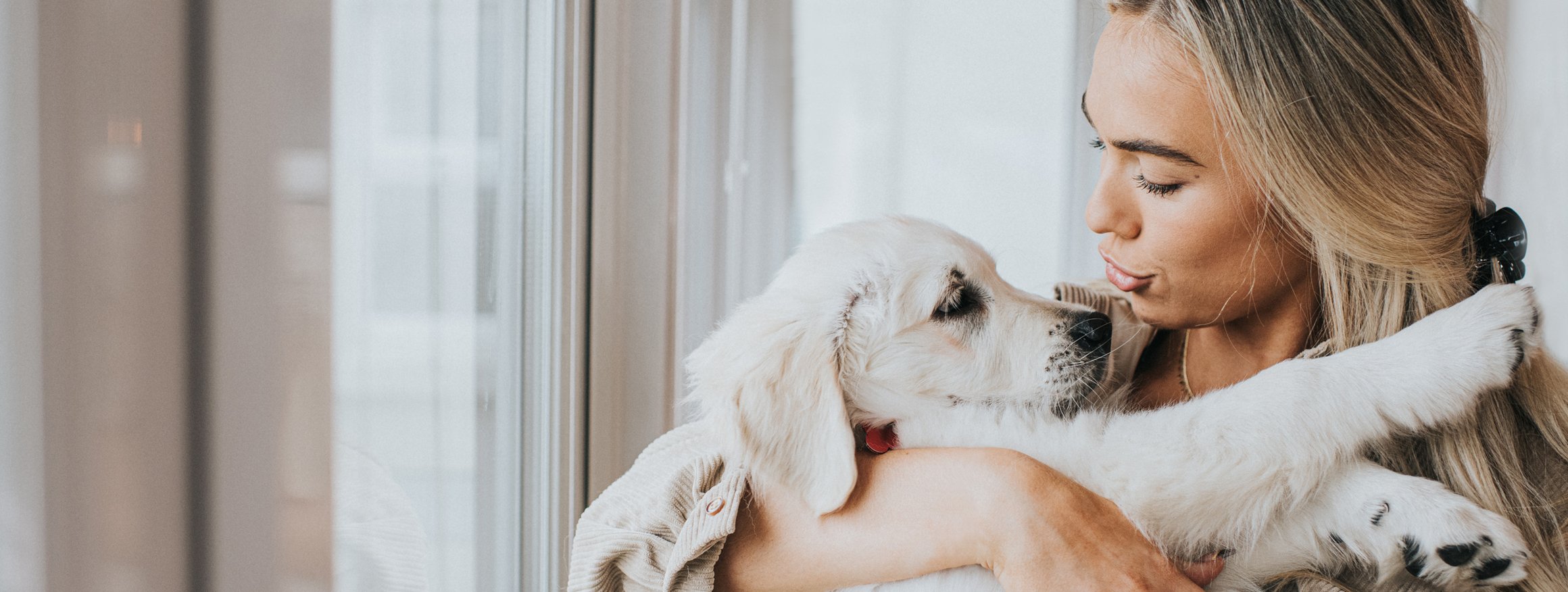 Eine junge Frau mit blonden langen Haaren hält fürsorglich einen jungen Hund im Arm und redet einfühlsam mit ihm