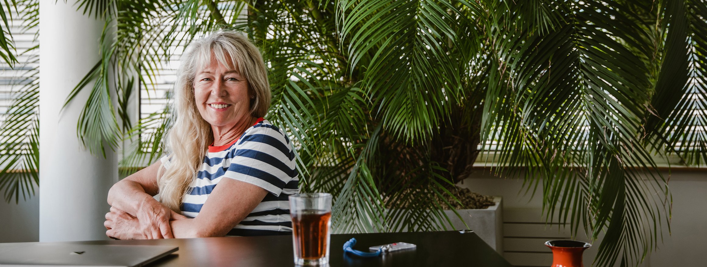 Die Stressforscherin Ulrike Ehlert sitzt entspannt in einem Zimmer an einem Tisch und lächelt, dahinter sind viele Palmen im Raum zu sehen