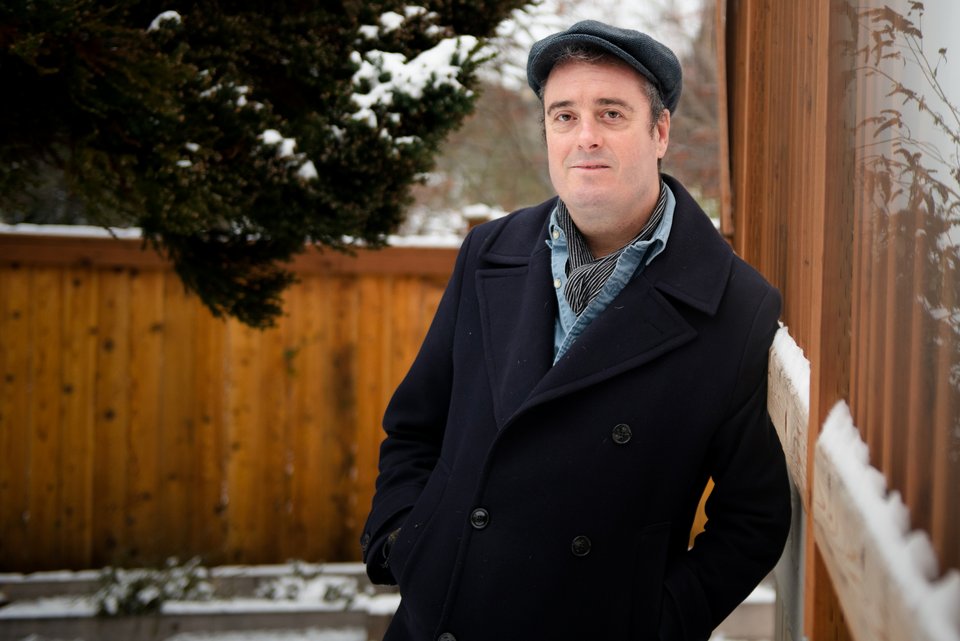 Der britische Psychologe Tim Lomas steht mit Mützen und Winterjacke auf einem Balkon, hinter ihm ein  schneebedeckter Garten mit Baum