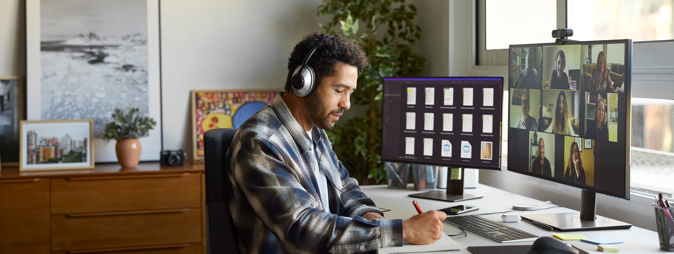 Ein Schwarzer Mann sitzt am Schreibtisch im Home-Office. Er trägt Kopfhörer und notiert etwas während eines digitalen Meetings.